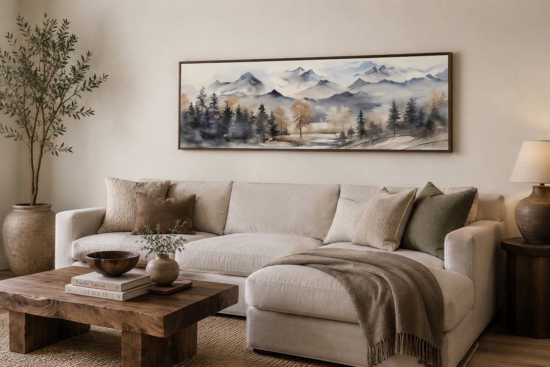 Horizontal framed canvas wall art of a watercolor mountain landscape in earth tones above a sofa.