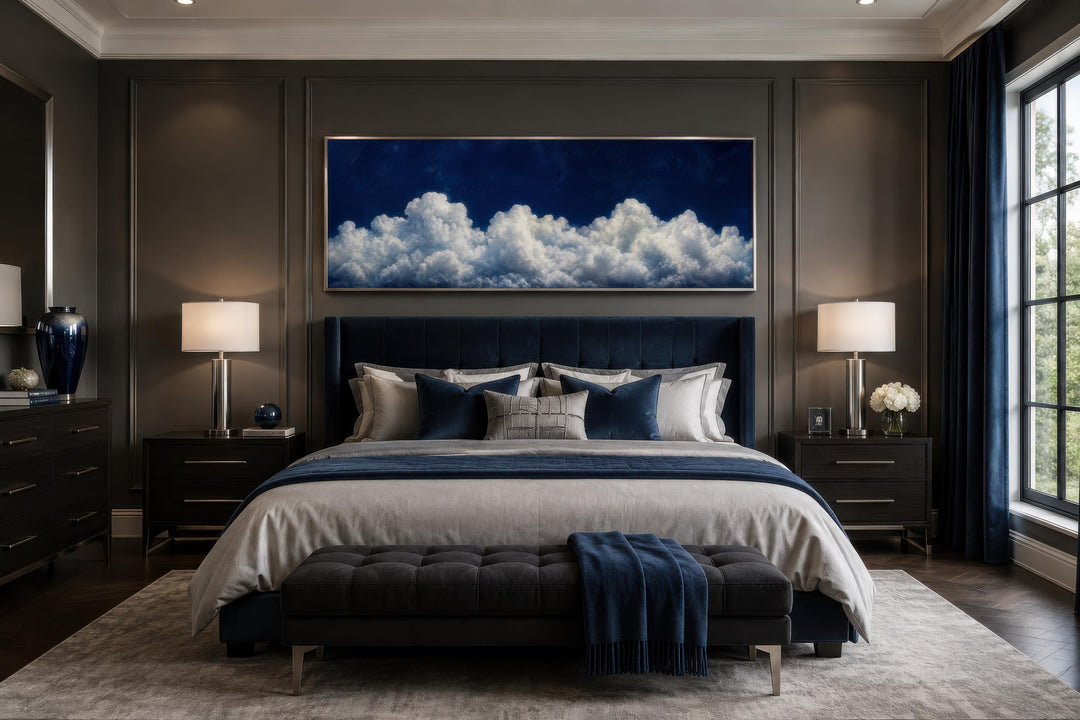 Long narrow canvas print of white clouds against a navy blue sky above a bed in a modern bedroom.