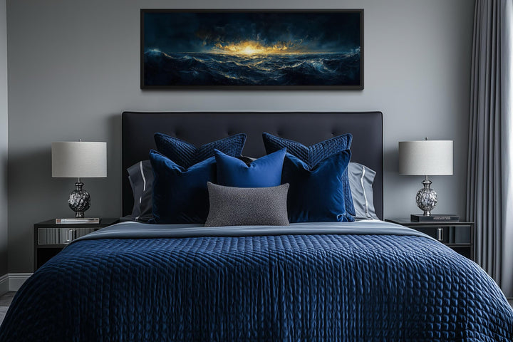 A horizontal navy and gold ocean sunset canvas print hangs above a modern bed with blue bedding.