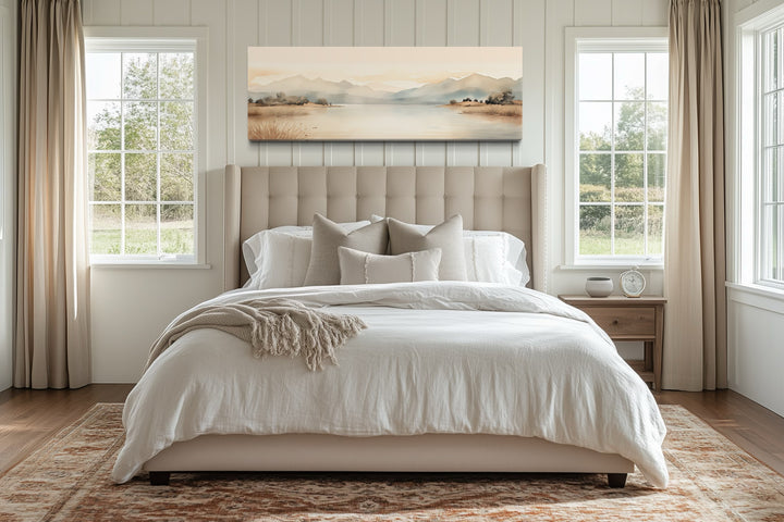 Long narrow framed canvas wall art of a beige mountain lake scene hanging above a bed.