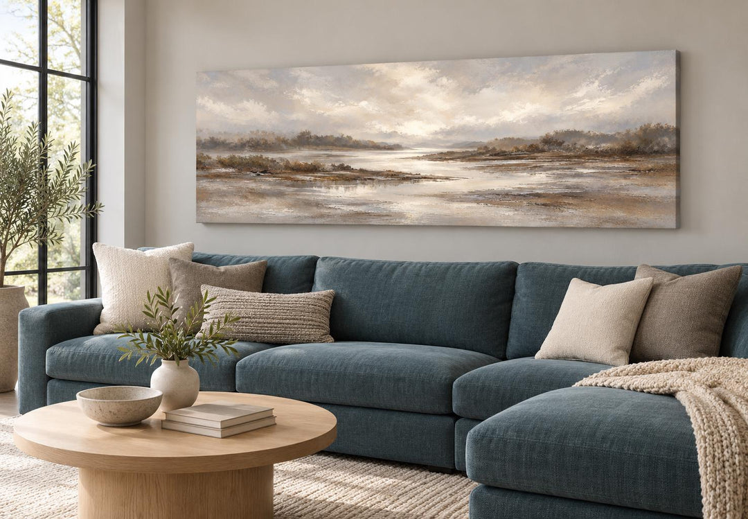 Neutral earth tones river landscape long horizontal canvas wall art above a blue sectional sofa.
