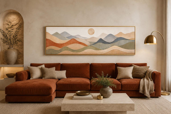 Panoramic canvas art with layered mountains in terracotta, blue, and taupe above a rust-toned sofa.