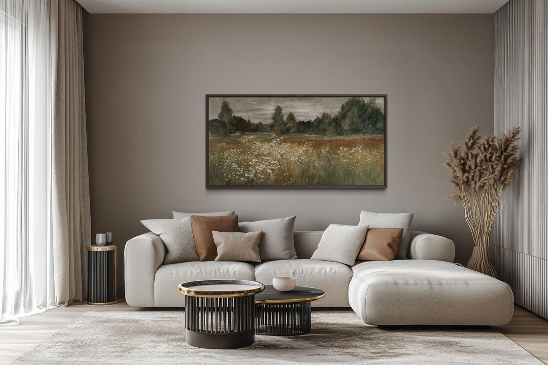 A framed horizontal canvas print of a neutral wildflower meadow landscape above a modern sofa.