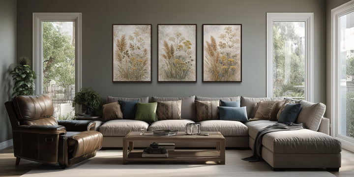 Set of three neutral wildflower botanical canvas prints in dark frames hanging above a sofa.