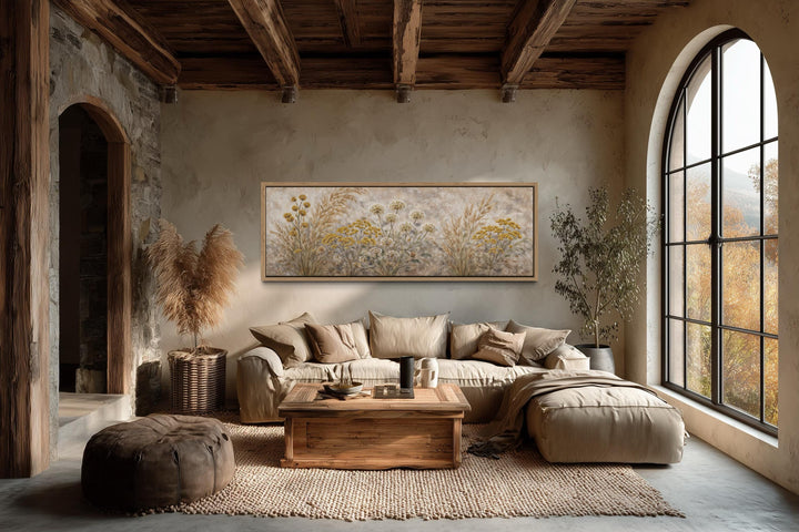 A long narrow canvas print of neutral wildflowers hangs above a sofa in a rustic, boho-style room.