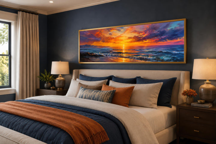 Ocean sunset wall art with a fiery orange and purple sky above a bed in a dark blue bedroom.