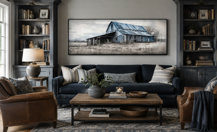 Long horizontal framed canvas wall art of an old rustic blue barn over a navy sofa in a living room.
