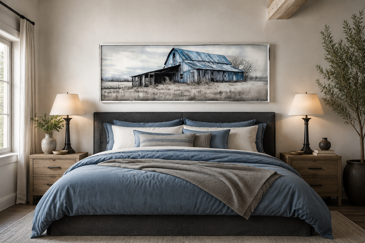 Long horizontal framed canvas wall art of a rustic blue barn over a bed in a farmhouse bedroom.