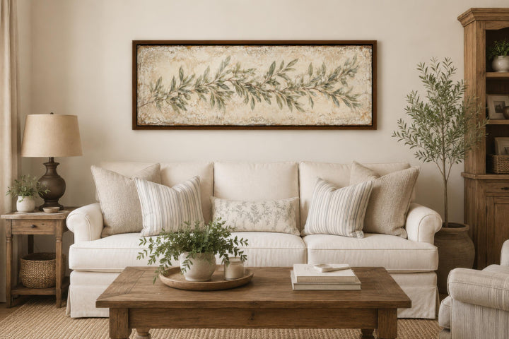 Long narrow canvas wall art featuring a sage green olive branch over a cream sofa in a living room.