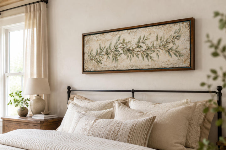 Long narrow canvas wall art of a sage green olive branch above a bed with neutral linens.