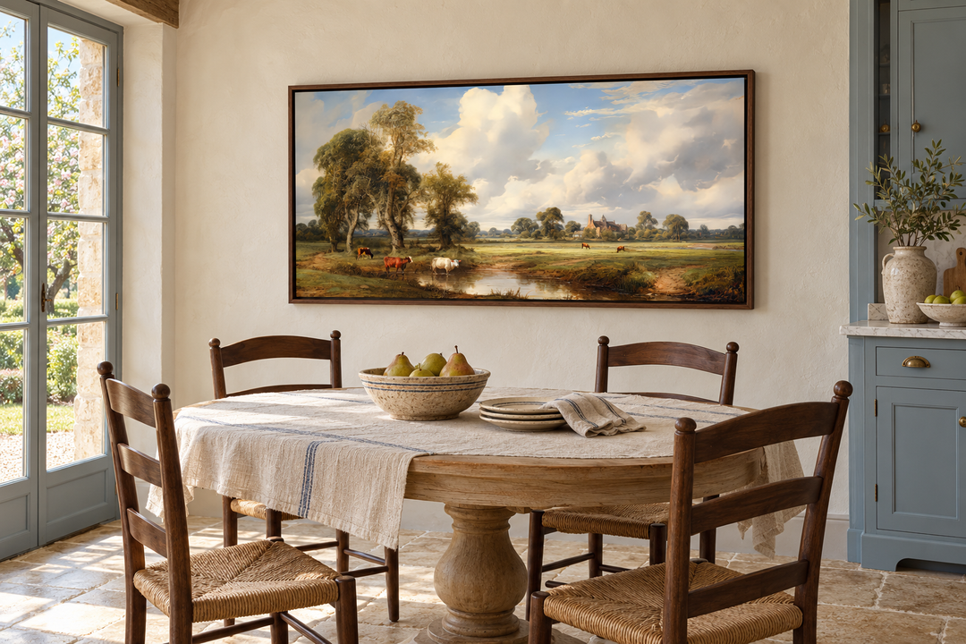Framed pastoral landscape painting of cows in a field above a rustic dining table with chairs.