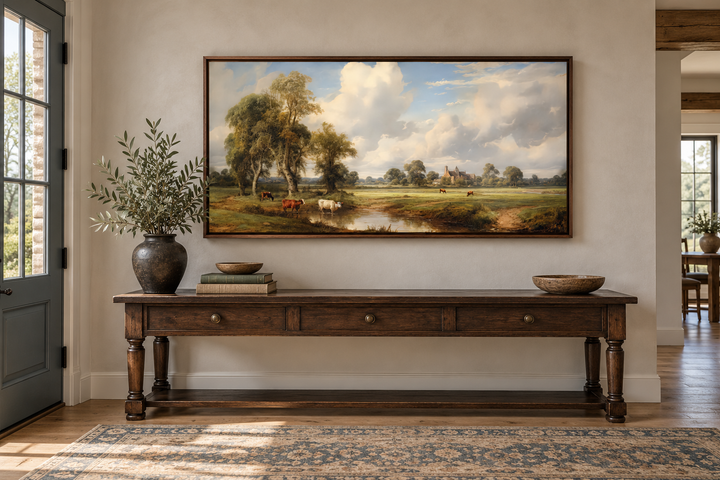 Framed antique pastoral landscape canvas with cows above a wooden console table in a farmhouse room.