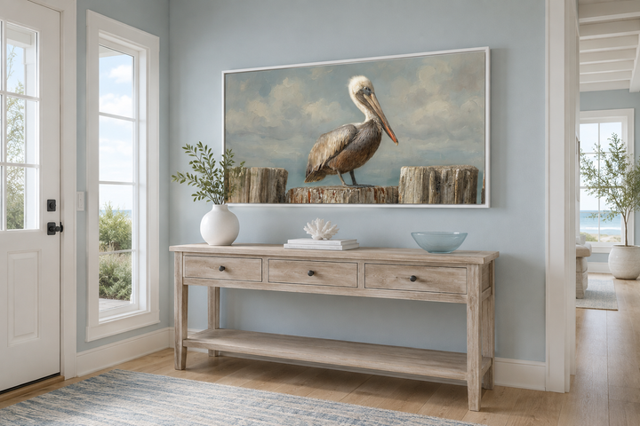 Horizontal canvas wall art of a pelican on wooden posts above a console table in a coastal room.