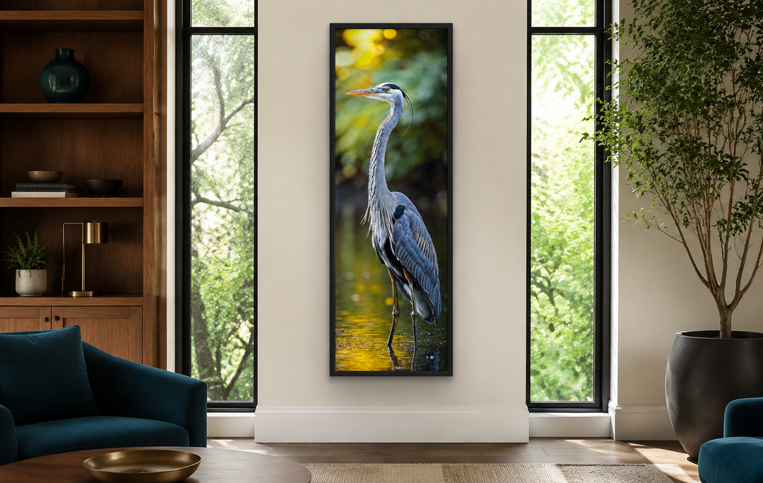 Vertical narrow wall art of a blue heron standing in water, framed in black on a neutral wall.