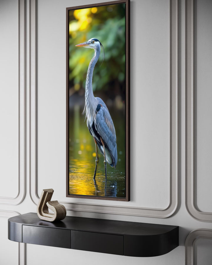 Vertical narrow wall art of a blue heron standing in water, framed in a dark wood frame.