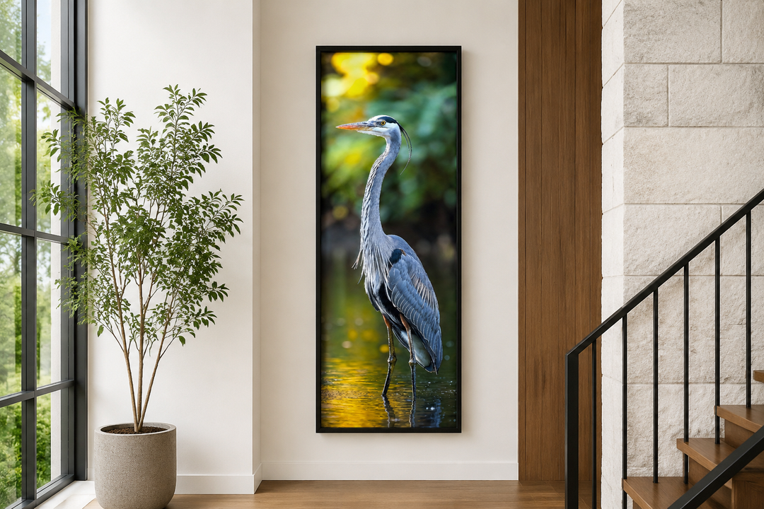 Vertical narrow wall art of a blue heron standing in water, framed in black on a white wall.