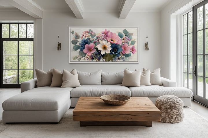 Horizontal framed canvas wall art of a blue and pink wildflower bouquet above a neutral sofa.