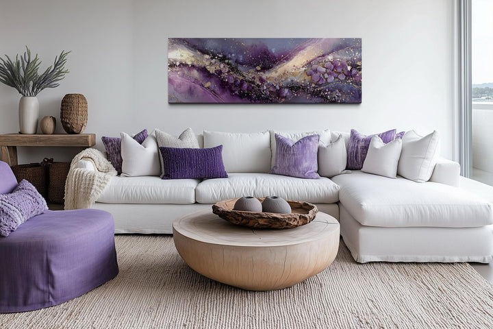 Purple abstract fluid landscape canvas print with gold accents hanging above a white sectional sofa.