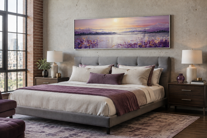 Long horizontal painting of a purple mountain lake sunset hanging above a grey upholstered bed.