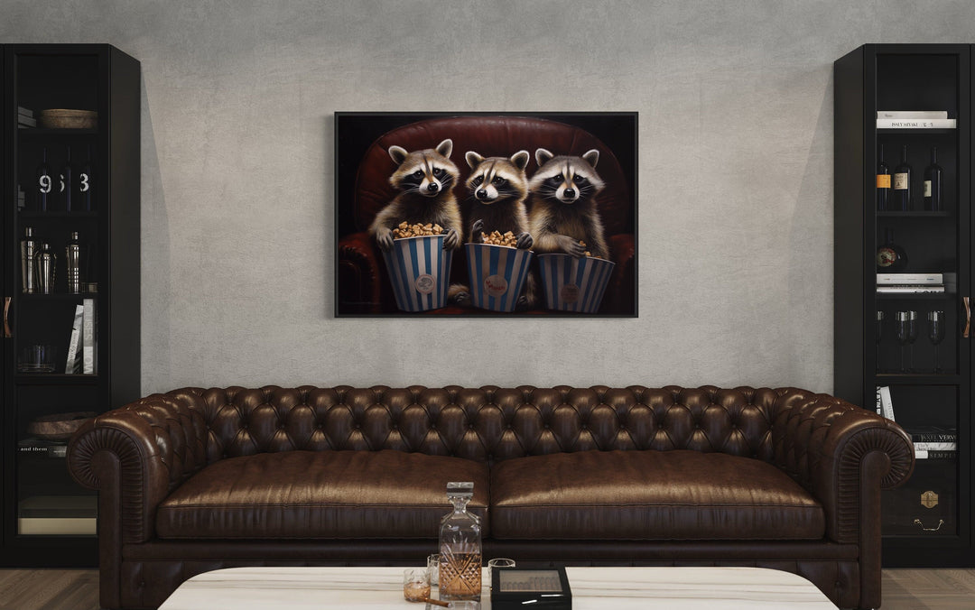 Framed canvas wall art of three raccoons eating popcorn in a theater above a brown leather sofa.