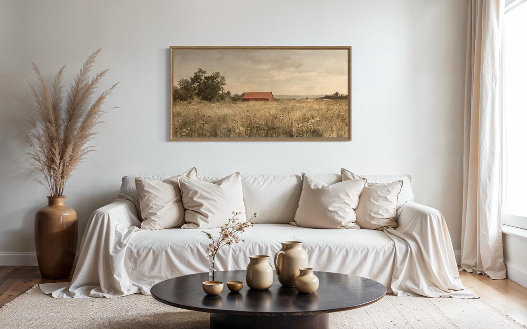 Red barn in a wildflower field landscape painting in a wood frame above a neutral sofa.