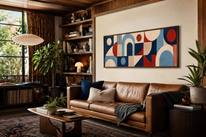 Long narrow Bauhaus canvas print with red, blue, and cream geometric shapes above a leather sofa.