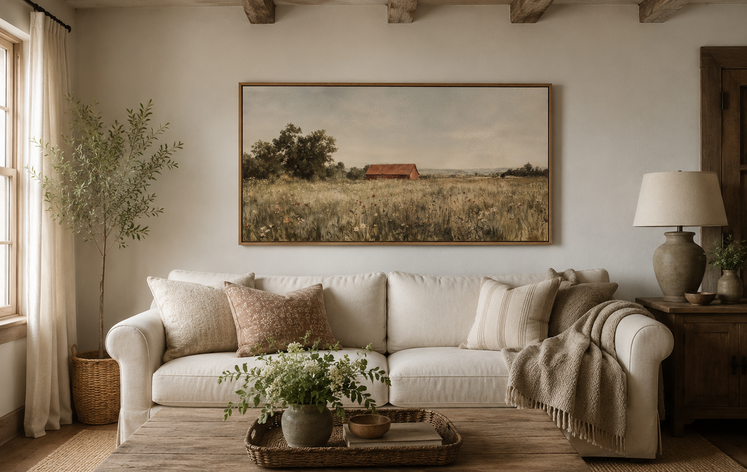 Red barn in a wildflower field landscape painting in a wooden frame above a white sofa.