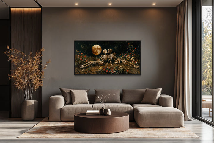 Romantic gothic wall art of two skeletons in a floral meadow under a full moon above a brown sofa.