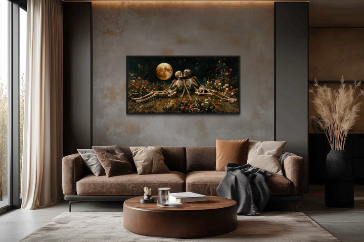 Eternal Love Skeletons wall art in a meadow under a full moon, hung above a brown sofa in a room.