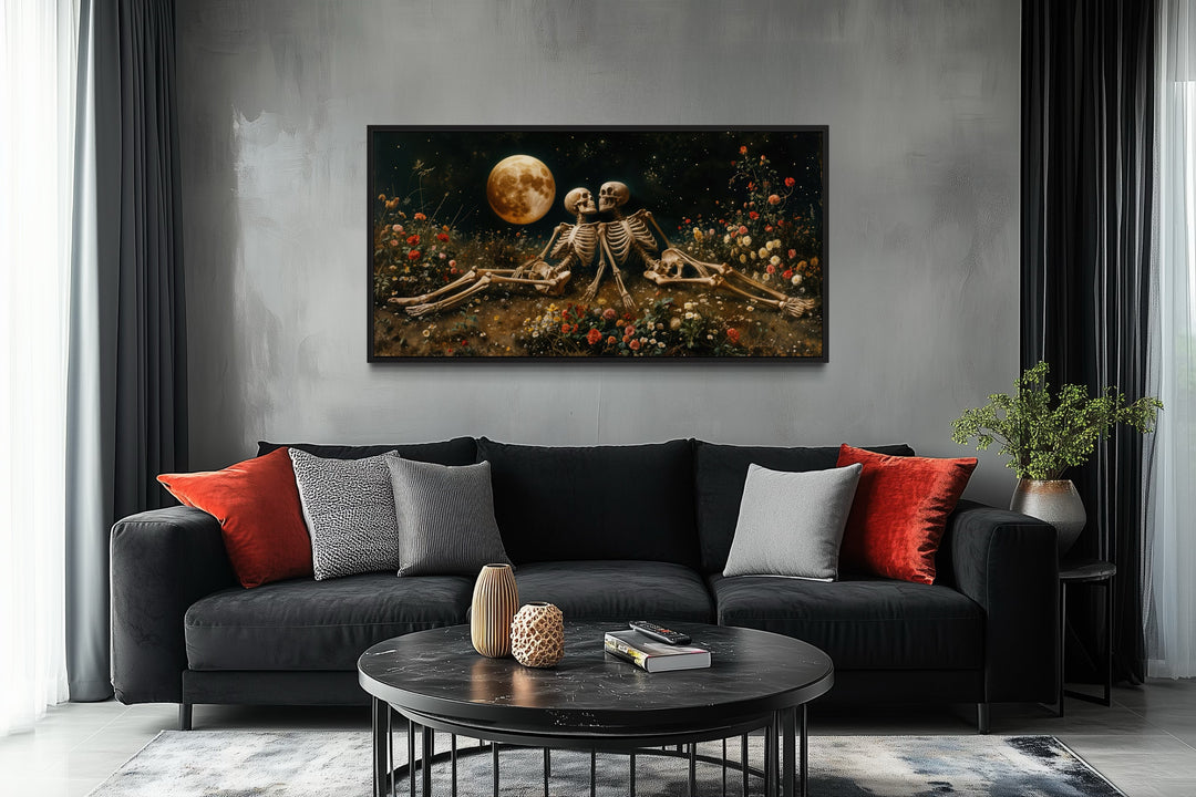 Romantic Gothic wall art of two skeletons in a floral meadow under a full moon above a black sofa.