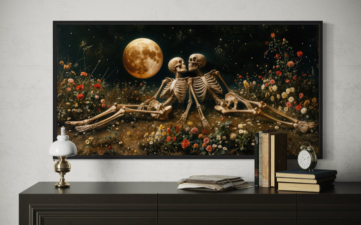 Framed gothic wall art of two skeletons in a flower meadow under a full moon above a dark dresser.