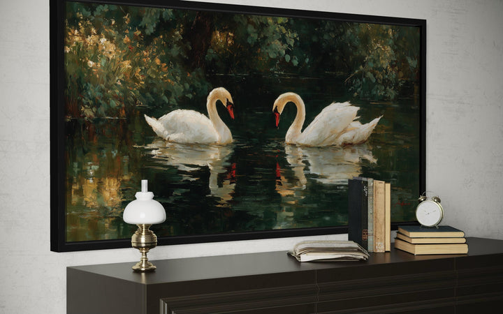 A framed canvas wall art of two white swans facing each other on a dark, reflective lake.