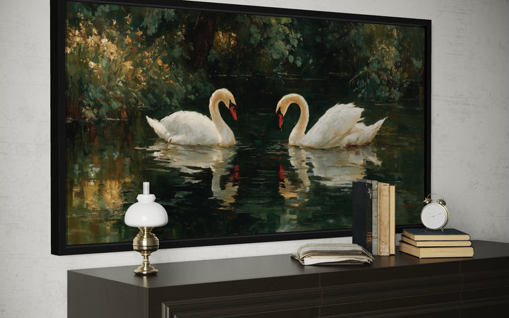 A framed canvas painting of two white swans on a dark lake, displayed above a dark wood dresser.