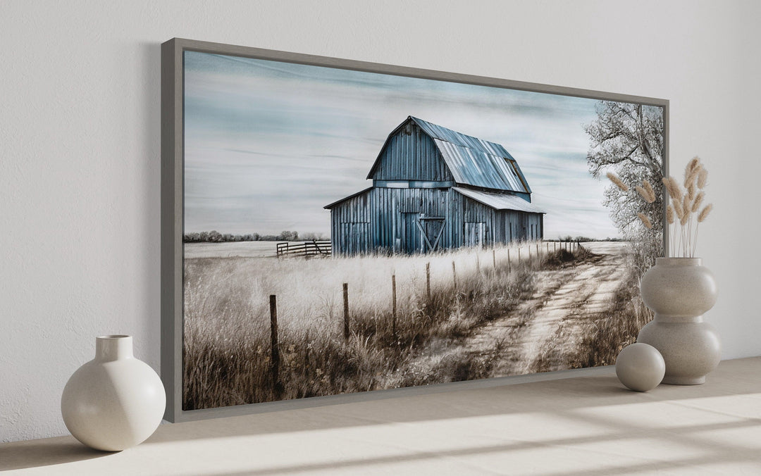 Rustic blue barn canvas painting in a gray frame, displayed on a shelf with decorative vases.