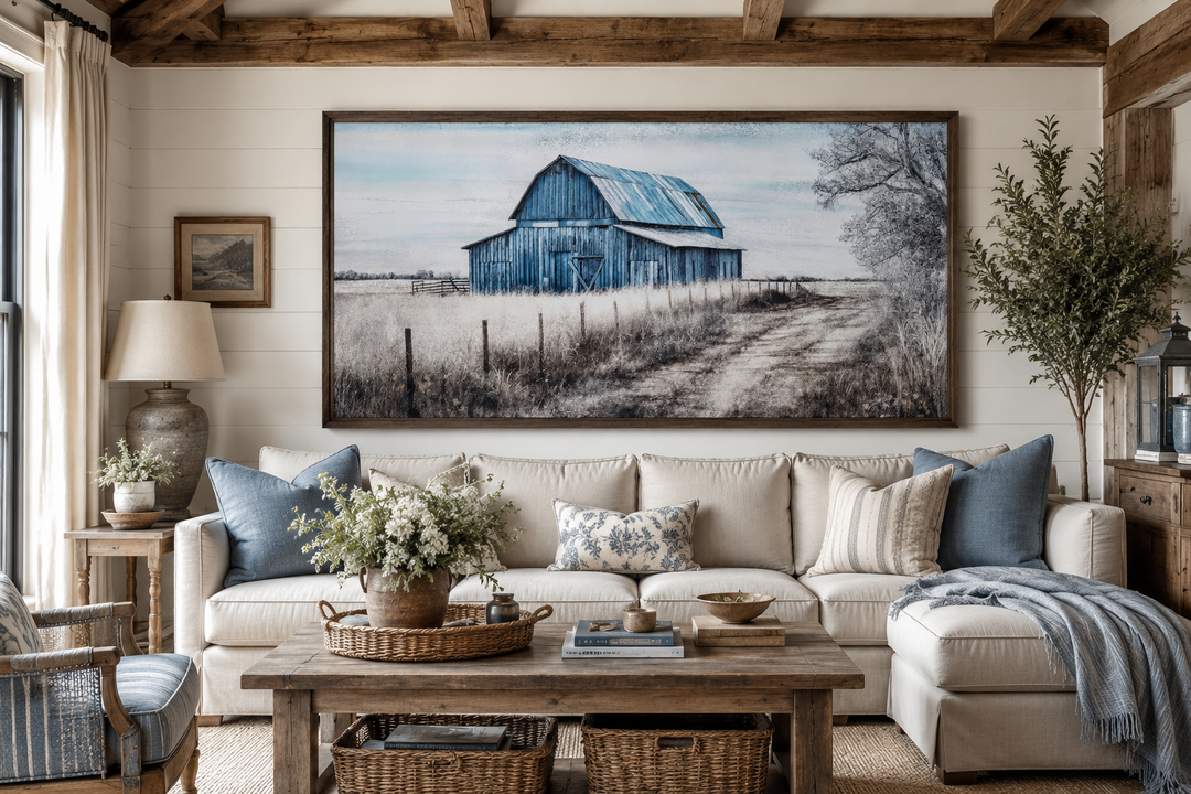Large framed canvas painting of a rustic blue barn in a field above a neutral farmhouse sofa.