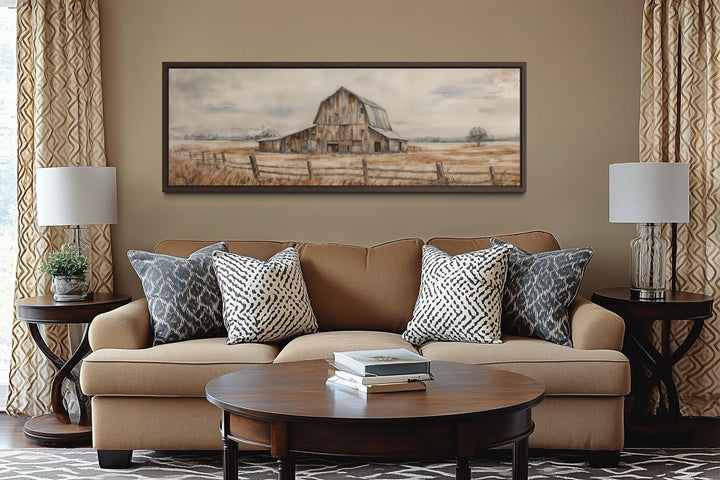 A long horizontal canvas print of a rustic barn in neutral tones hangs above a tan sofa.