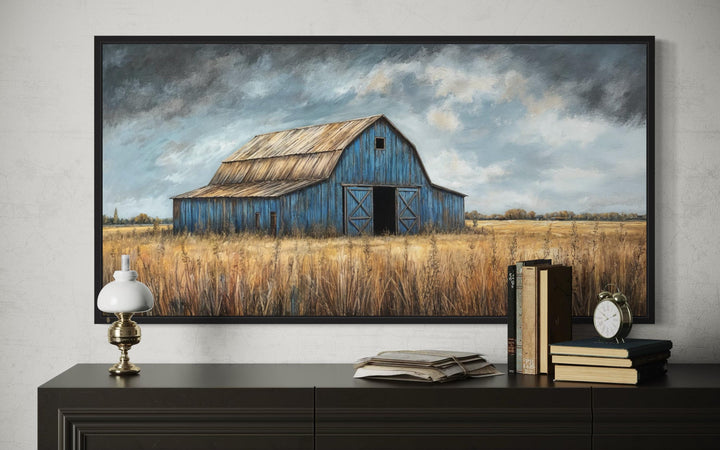 Rustic blue barn framed canvas wall art above a dark wood dresser with vintage books and a lamp.
