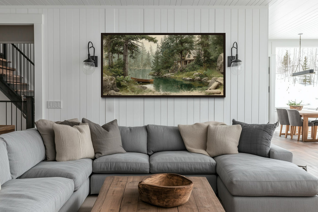 Framed panoramic painting of a wooden boat on a lake by a cabin, hung above a gray sectional sofa.