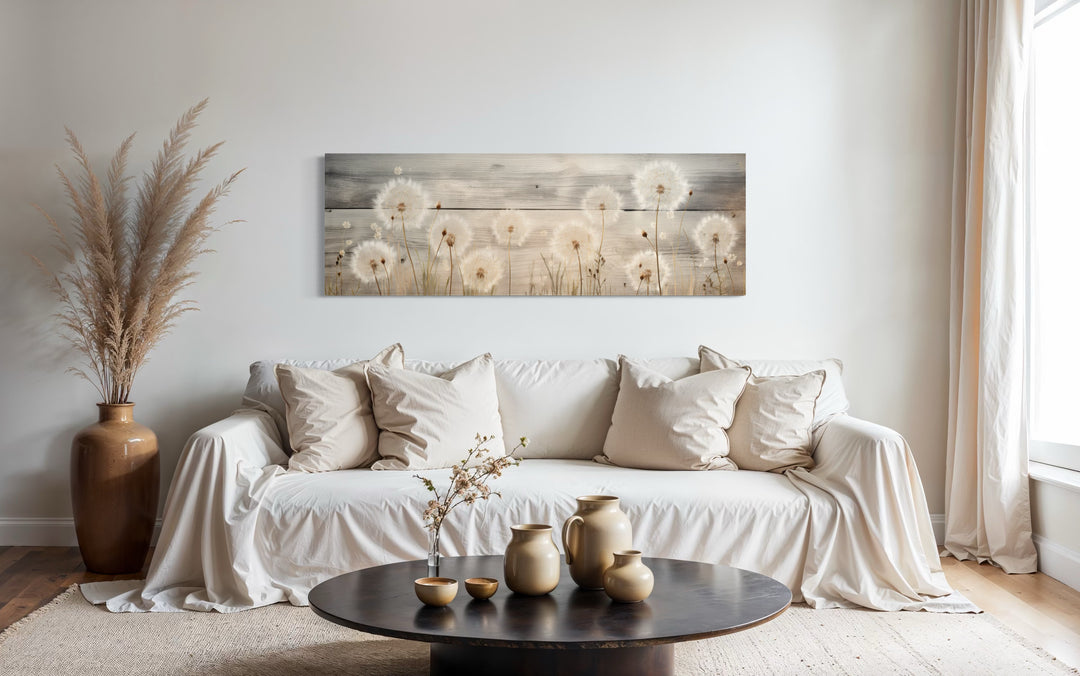 Rustic horizontal dandelion painting on wood hanging above a white sofa in a neutral living room.