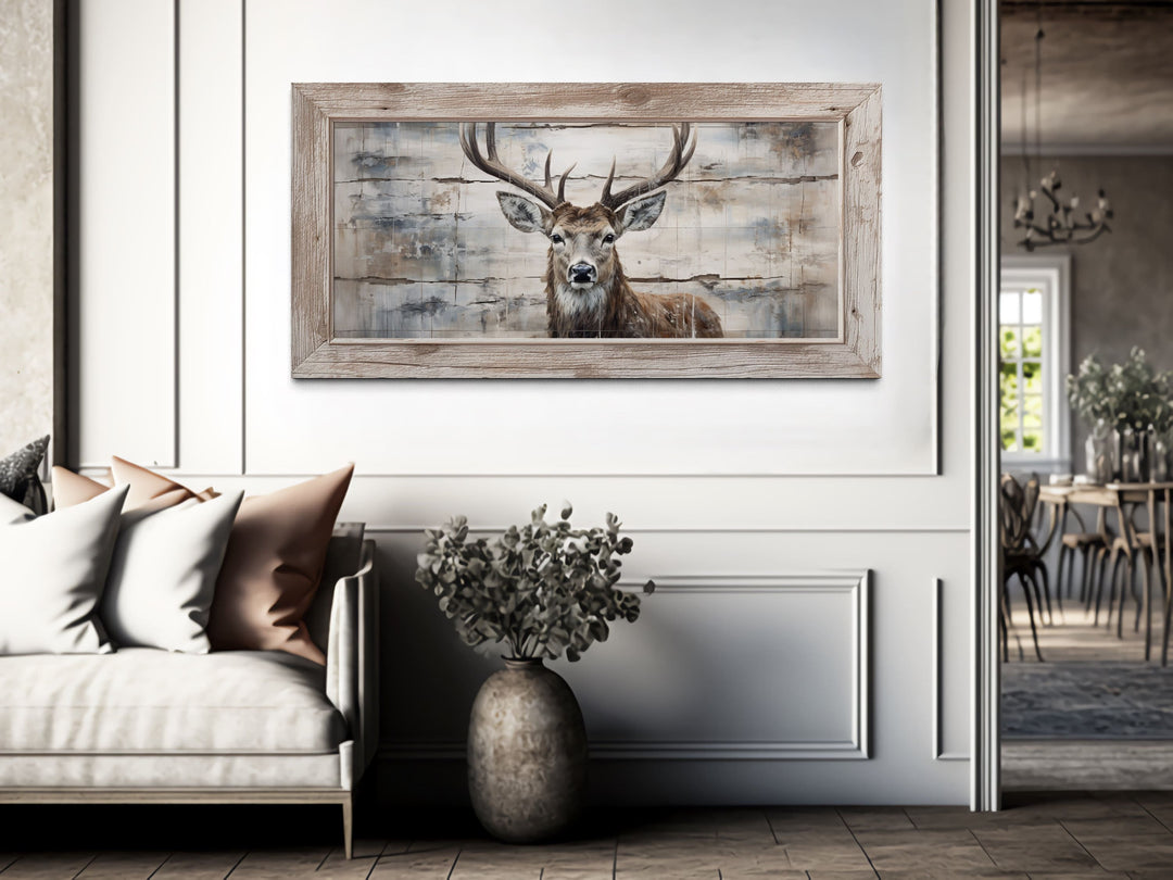 Rustic deer painting on wood-textured canvas in a light wood frame above a sofa in a neutral room.