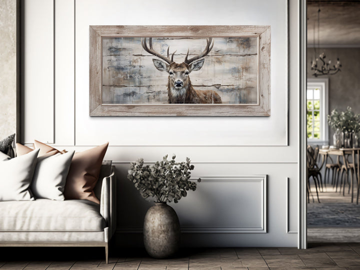 Rustic deer painting on wood-textured canvas in a light wood frame above a sofa in a neutral room.