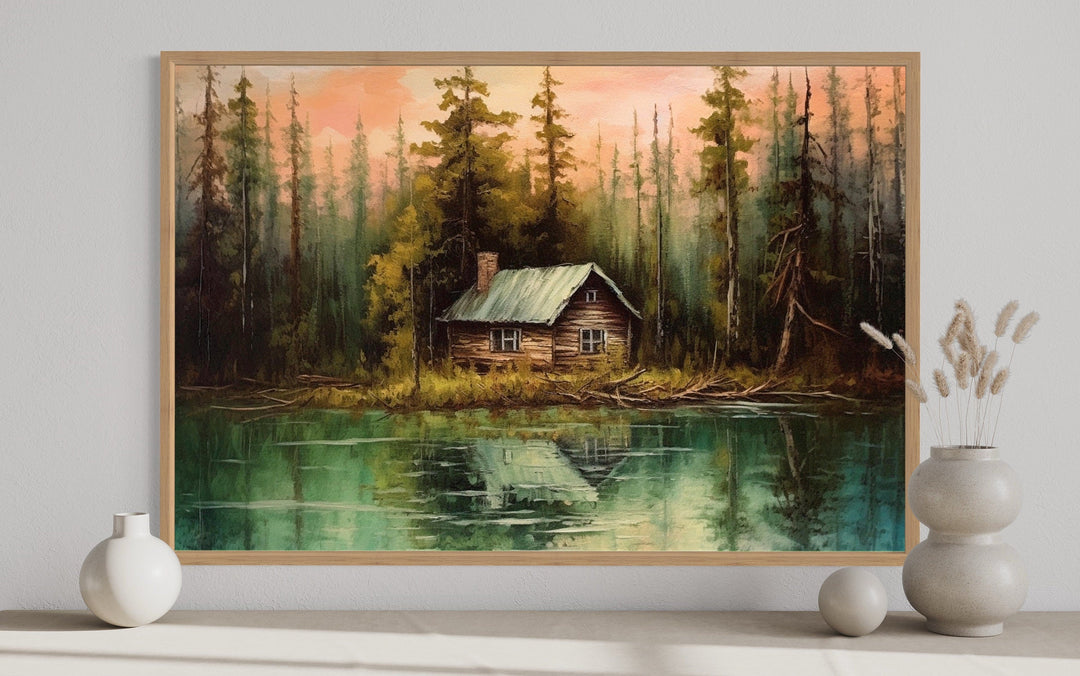 Rustic framed canvas wall art of a cabin by a lake, displayed on a shelf with decorative vases.