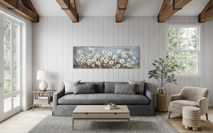 Rustic white daisy wildflowers painted on a long horizontal wood-style canvas above a gray sofa.