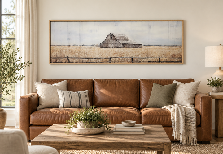 Rustic old barn painting on long narrow framed canvas wall art above a brown leather sofa.