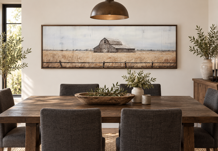Rustic old barn painting on long narrow framed canvas wall art above a wooden dining table.
