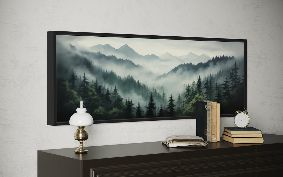 Sage green foggy pine forest and mountain landscape wall art in a black frame above a dark dresser.