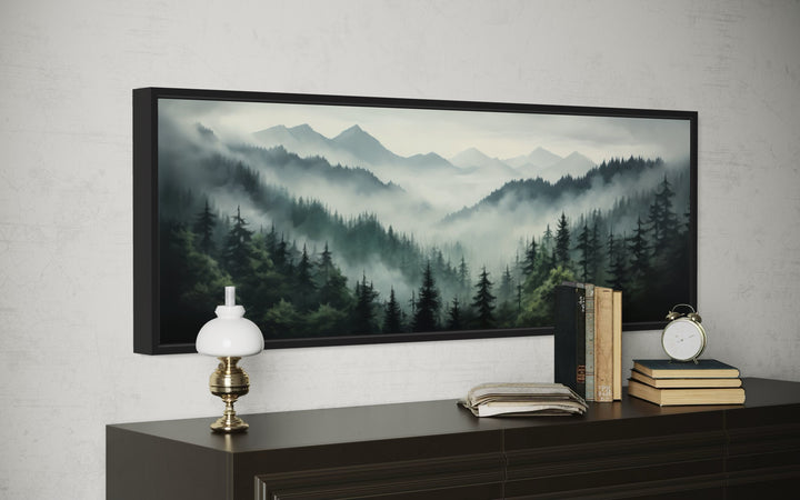 Sage green foggy pine forest and mountain landscape wall art in a black frame above a dark dresser.