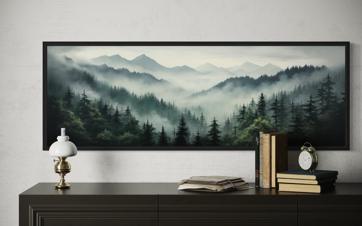 Sage green foggy pine forest and mountain landscape wall art in a black frame above a dark dresser.