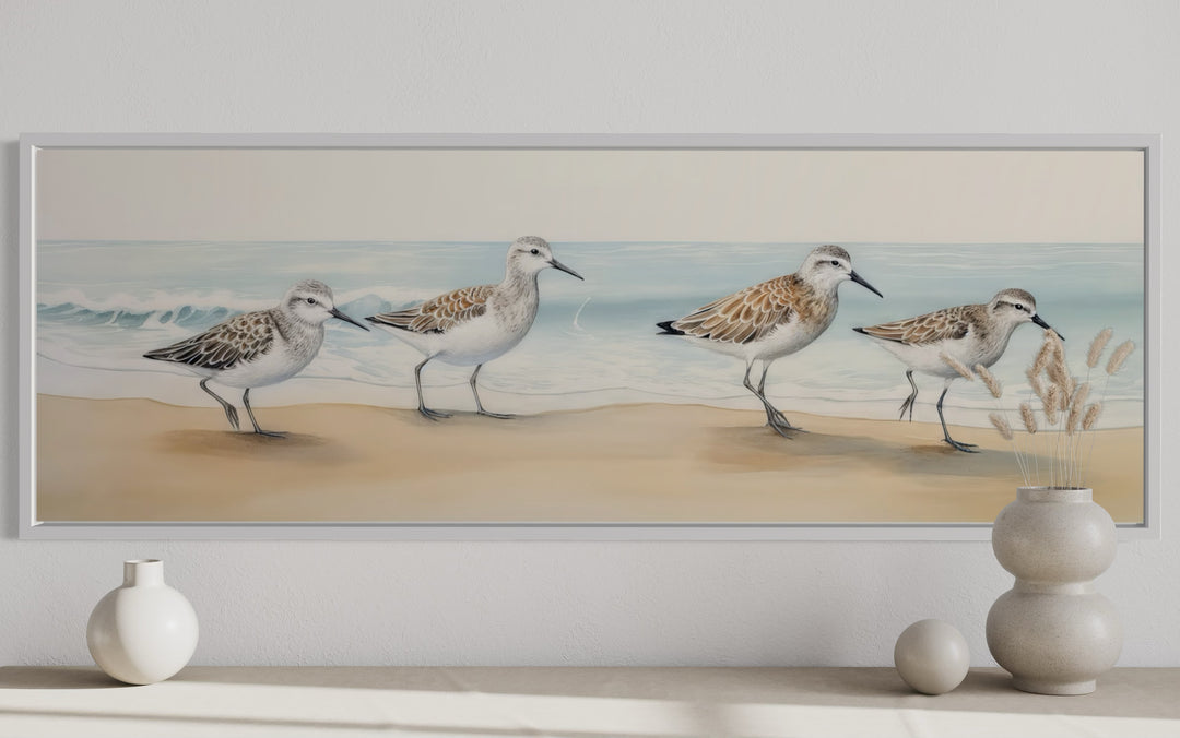 Horizontal framed canvas wall art of four sandpipers walking on a sandy beach by the ocean.