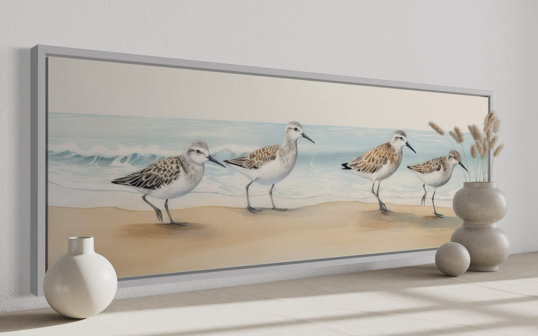 Horizontal framed canvas wall art of four sandpipers walking on a sandy beach by the ocean.